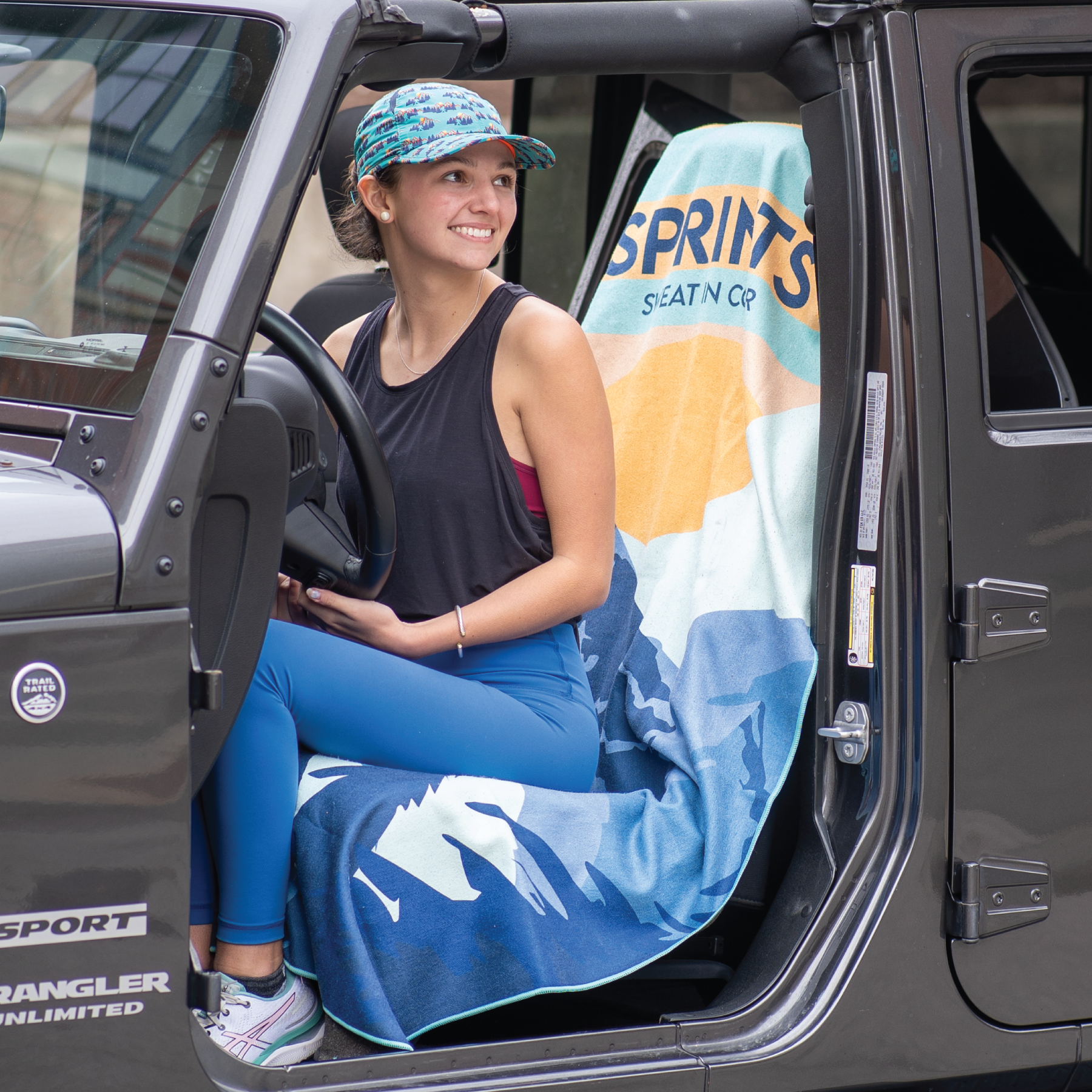 Woman sitting on towel covering car seat of jeep, with image of Sasquatch character against mountains and tress in blue and orange and teal color.