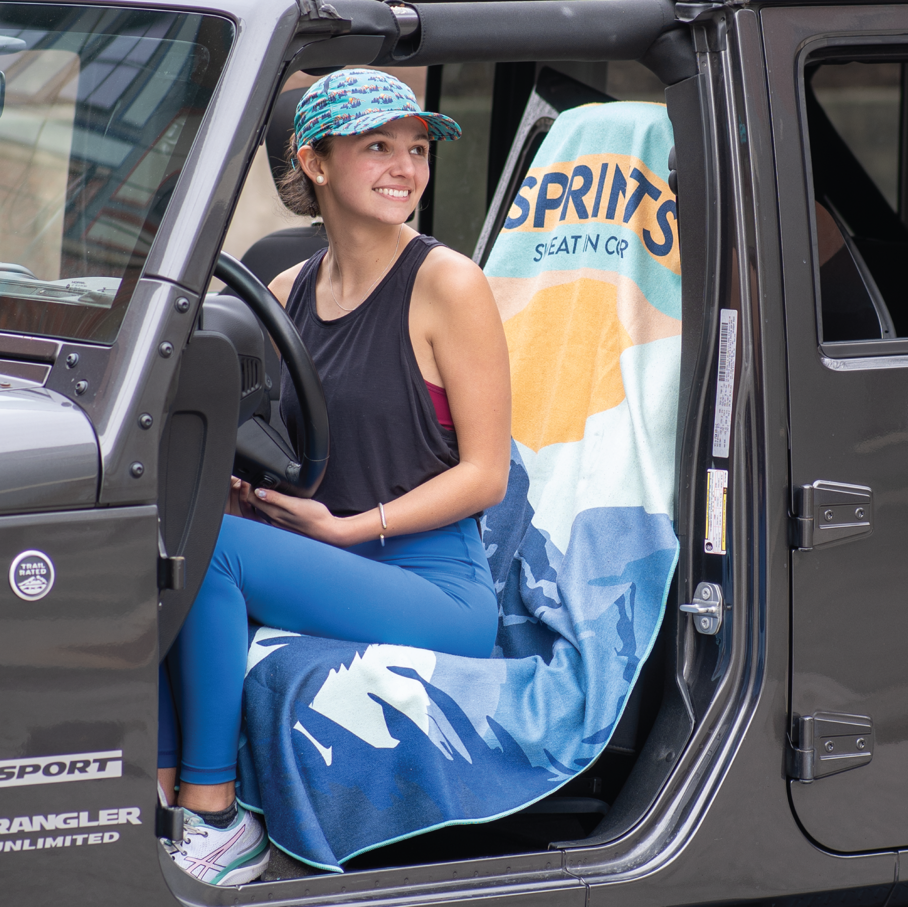 Woman sitting on towel covering car seat of jeep, with image of Sasquatch character against mountains and tress in blue and orange and teal color.