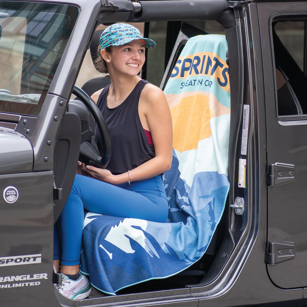 Woman sitting on towel covering car seat of jeep, with image of Sasquatch character against mountains and tress in blue and orange and teal color. 