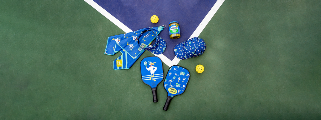 Pickleball paddles, hat, visor and mini towel with vlasic pickles repeating pattern on each item laying on a pickleball court