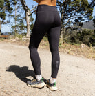 A woman standing outdoors on a dirt path wearing black high-waisted athletic leggings and a maroon sports bra, photographed from behind with trees and blue sky in the background.