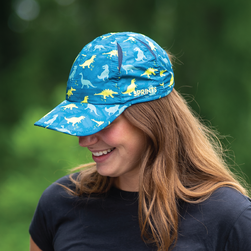 Picture of girl looking down and smiling, she is wearing a blue and green hat with dinosaurs on it
