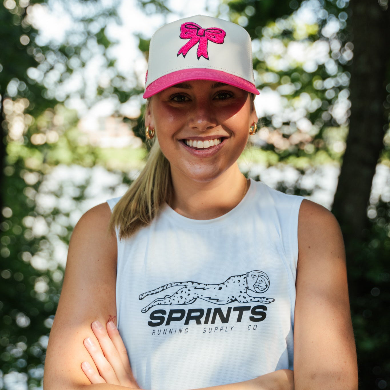 A smiling woman outdoors wearing a white sleeveless athletic tank top with a black Sprints logo and a white trucker cap with a pink bow. Trees and sunlight are blurred in the background.