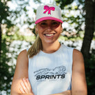 A smiling woman outdoors wearing a white sleeveless athletic tank top with a black Sprints logo and a white trucker cap with a pink bow. Trees and sunlight are blurred in the background.