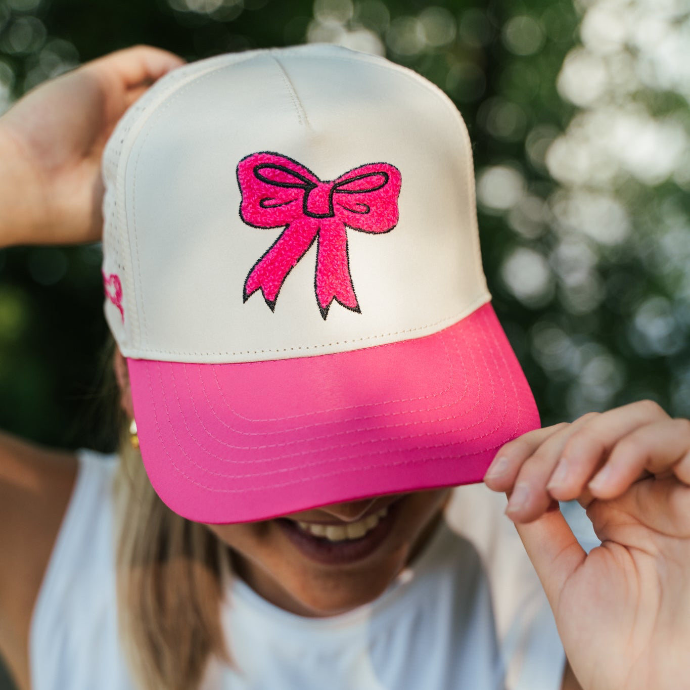 A person outdoors wearing a white and bright pink trucker-style cap with a pink bow graphic on the front, partially covering their face. The person is also wearing a white sleeveless shirt with 'SPRINTS' printed on the front.
