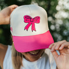 A person outdoors wearing a white and bright pink trucker-style cap with a pink bow graphic on the front, partially covering their face. The person is also wearing a white sleeveless shirt with 'SPRINTS' printed on the front.