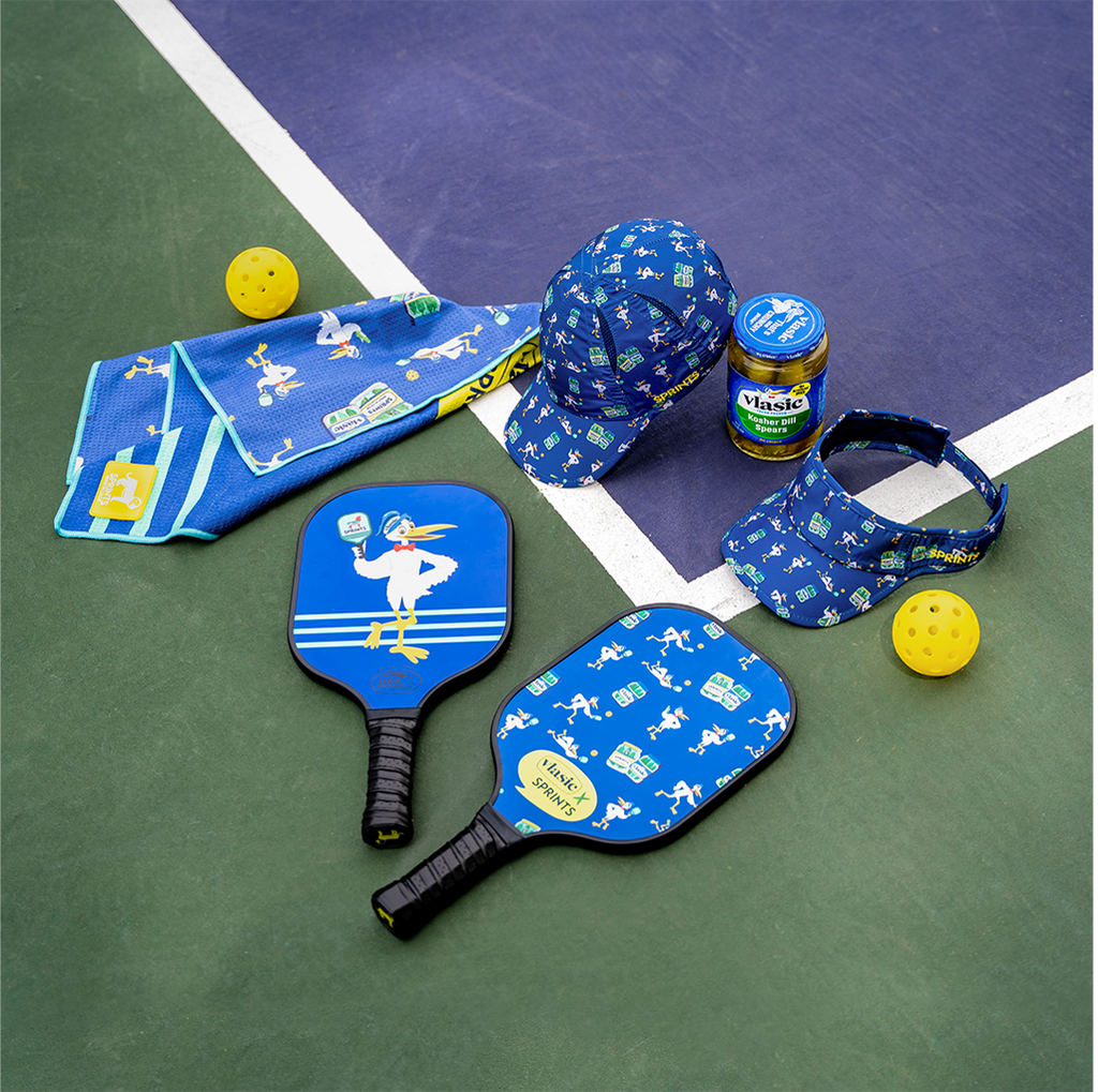 Pickleball paddles, hat, visor and mini towel with vlasic pickles repeating pattern on each item laying on a pickleball court