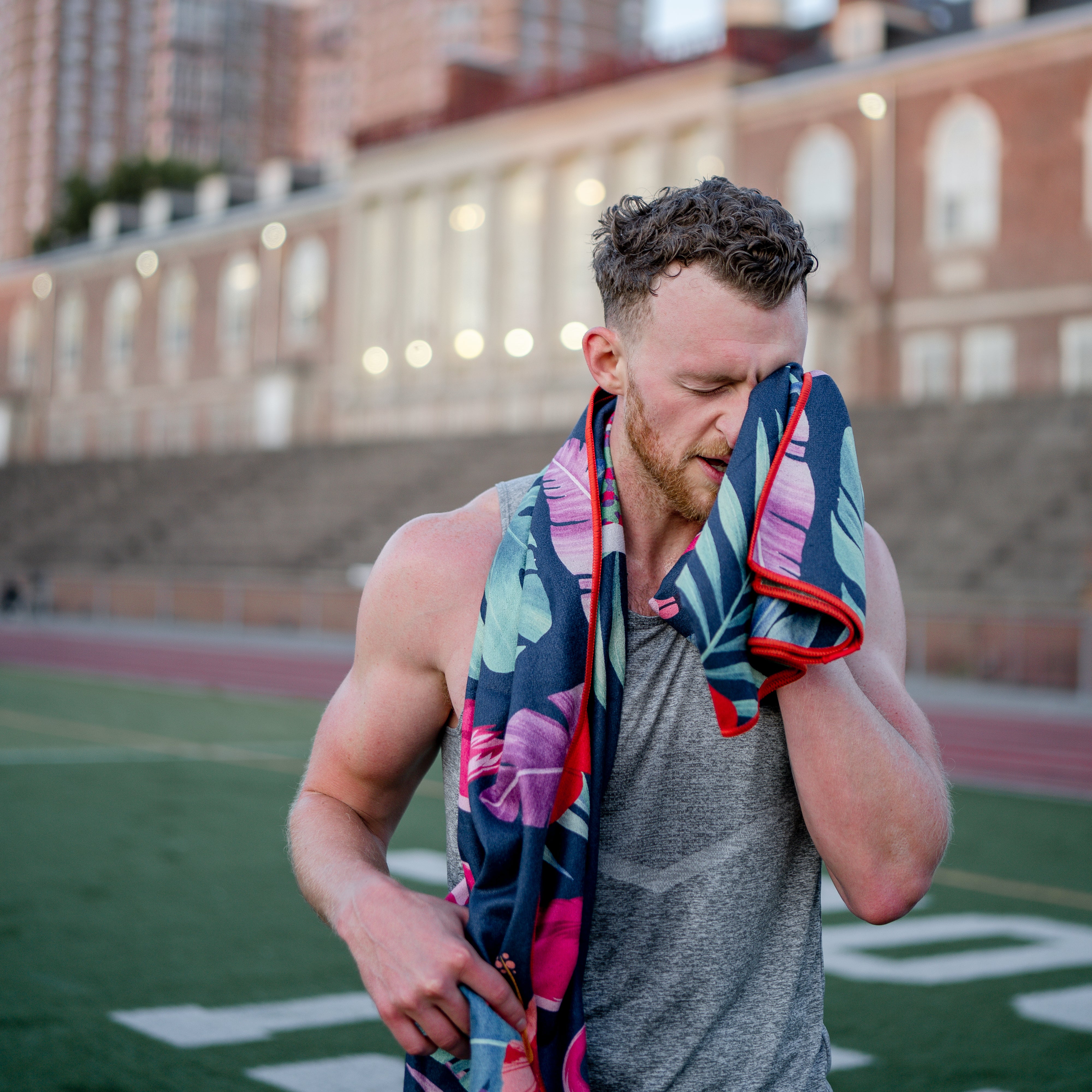 Towel with pattern of pink flamingos against tropical leaves in dark blue and green background being used to wipe seat off mans face at a track