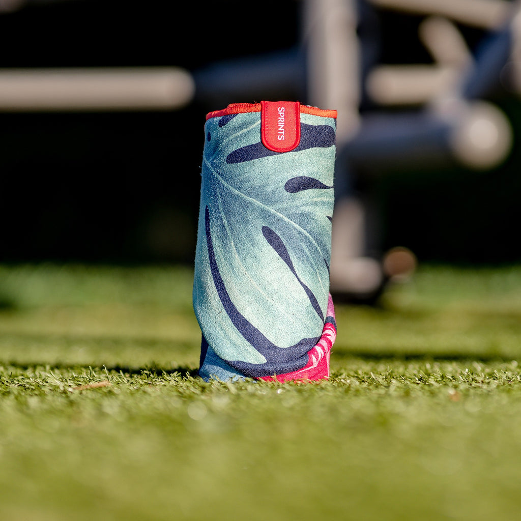 Towel with pattern of pink flamingos against tropical leaves in dark blue and green background rolled up and sitting on the grass. 