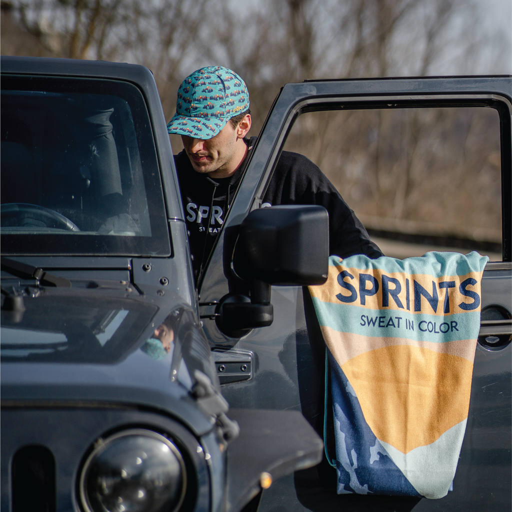Towel with image of Sasquatch character against mountains and tress in blue and orange and teal color sitting over driverside door of jeep while man gets in the car. 
